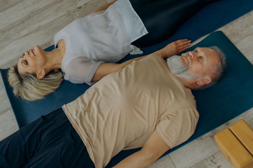 Elderly couple lying on yoga mats in a peaceful indoor setting, embracing togetherness.